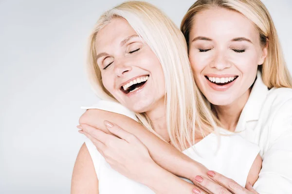 Lächelnde blonde reife Mutter und kleine Tochter in ganz weißer Kleidung umarmen sich mit geschlossenen Augen isoliert auf grau — Stockfoto