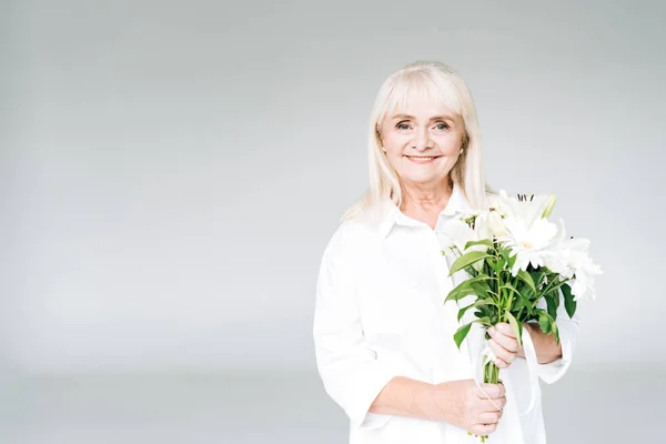 Happy blonde senior woman in white clothes with flowers isolated on grey with copy space — Stock Photo