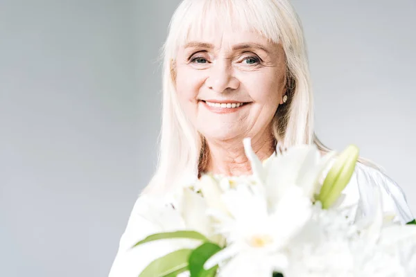 Happy blonde senior woman in white clothes holding flowers isolated on grey — Stock Photo