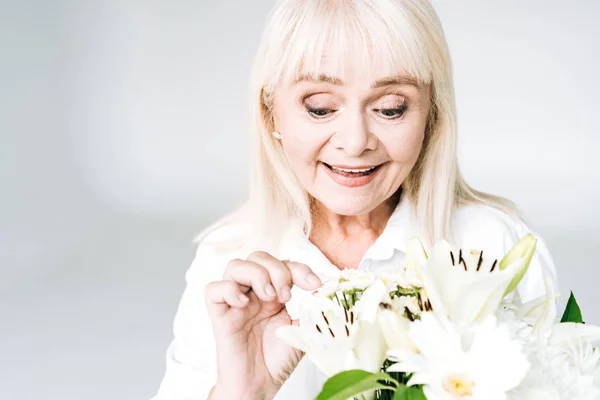 Exited blonde senior woman in white clothes with flowers isolated on grey — Stock Photo