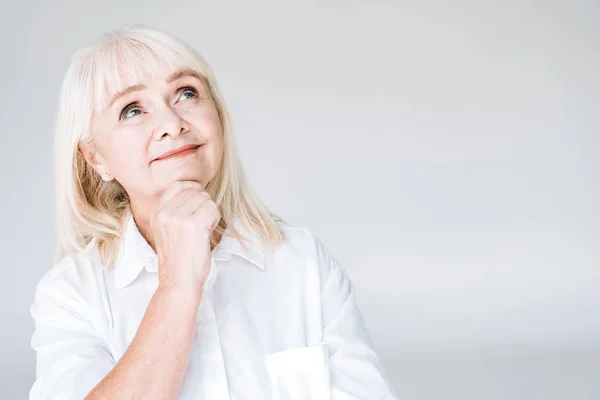 Heureuse blonde rêveuse femme âgée en vêtements blancs regardant loin isolé sur gris — Photo de stock