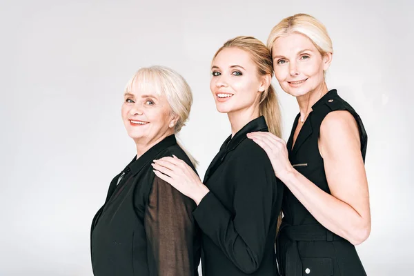 Vista lateral de las mujeres rubias de moda de tres generaciones en trajes negros totales aislados en gris - foto de stock