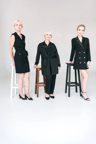 Full length view of fashionable three-generation blonde women in total black outfits near chairs isolated on grey — Stock Photo
