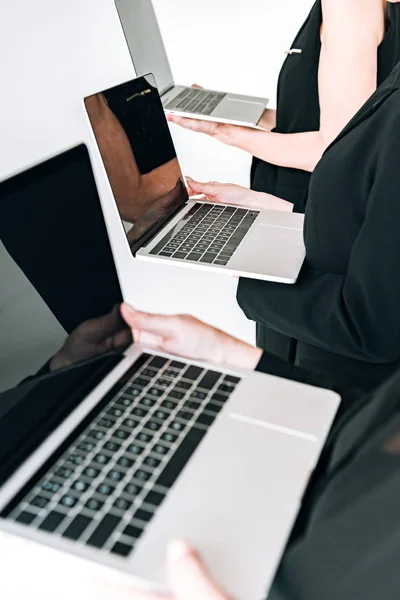 Vista recortada de las empresarias de tres generaciones en trajes negros totales que sostienen computadoras portátiles con pantallas en blanco aisladas en gris - foto de stock