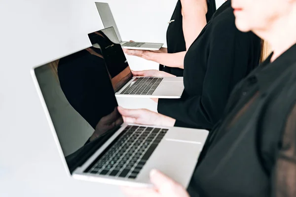 Vista parcial de las empresarias de tres generaciones en trajes negros totales que sostienen computadoras portátiles con pantallas en blanco aisladas en gris - foto de stock