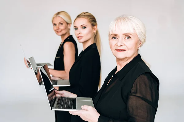 Vista lateral de las mujeres de negocios rubias de moda de tres generaciones en trajes negros totales que sostienen computadoras portátiles aisladas en gris - foto de stock