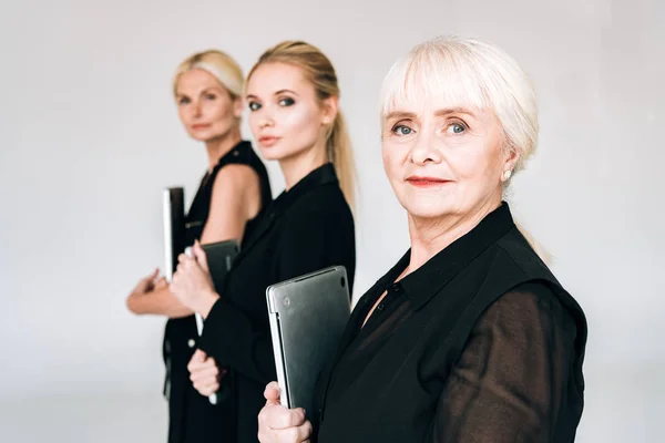 Enfoque selectivo de tres generaciones de mujeres de negocios rubias en trajes negros totales que sostienen computadoras portátiles aisladas en gris - foto de stock