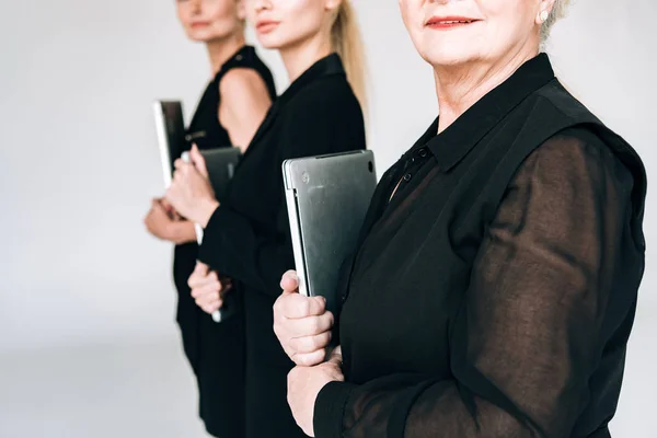 Vista ritagliata di donne d'affari bionde di tre generazioni in abiti neri totali che tengono i computer portatili isolati su grigio — Foto stock