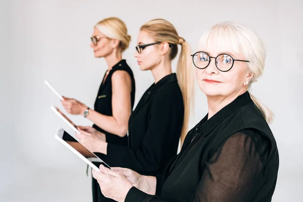 Mujeres de negocios rubias de moda de tres generaciones en trajes negros totales y gafas que sostienen tabletas digitales aisladas en gris - foto de stock