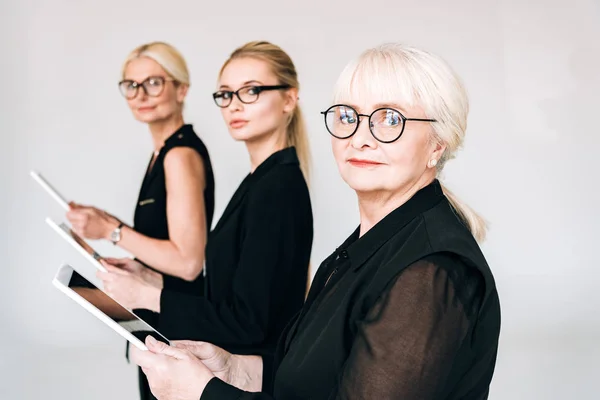 Femmes d'affaires blondes de trois générations en tenue noire totale et lunettes tenant des tablettes numériques isolées sur gris — Photo de stock