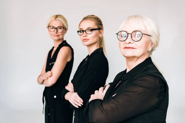 Selective focus of fashionable three-generation blonde businesswomen in total black outfits and glasses isolated on grey — Stock Photo
