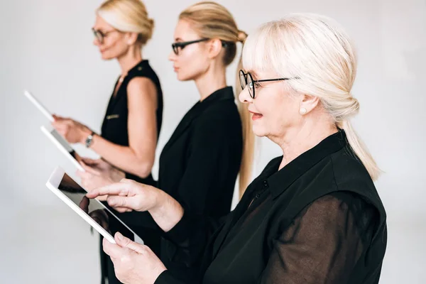 Mujeres de negocios rubias de moda de tres generaciones en trajes y gafas negros totales utilizando tabletas digitales aisladas en gris - foto de stock