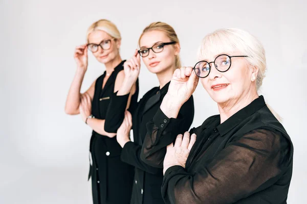 Mulheres de negócios loira de três gerações na moda em roupas pretas totais tocando óculos isolados em cinza — Fotografia de Stock