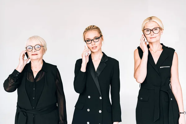 Mujeres de negocios de moda de tres generaciones en trajes negros totales y gafas hablando en teléfonos inteligentes aislados en gris - foto de stock
