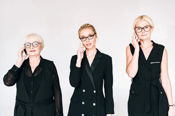 Modische Geschäftsfrauen der dritten Generation in komplett schwarzen Outfits und Brille, die auf Smartphones sprechen, isoliert auf grau — Stockfoto