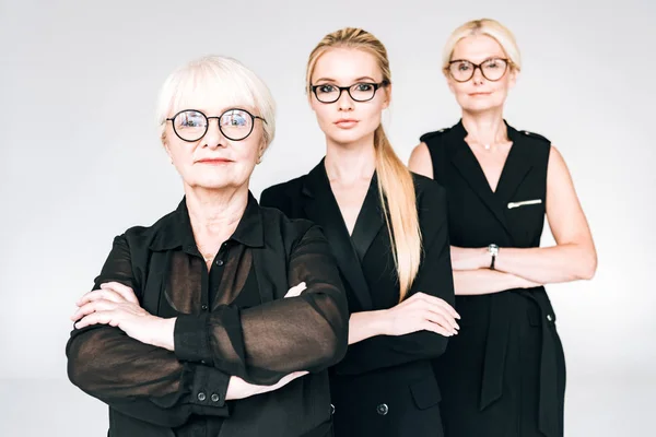 Mujeres de negocios rubias de moda de tres generaciones en gafas con brazos cruzados aislados en gris - foto de stock