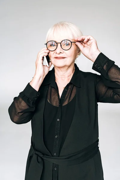 Senior businesswoman in black outfit and glasses talking on smartphone isolated on grey — Stock Photo