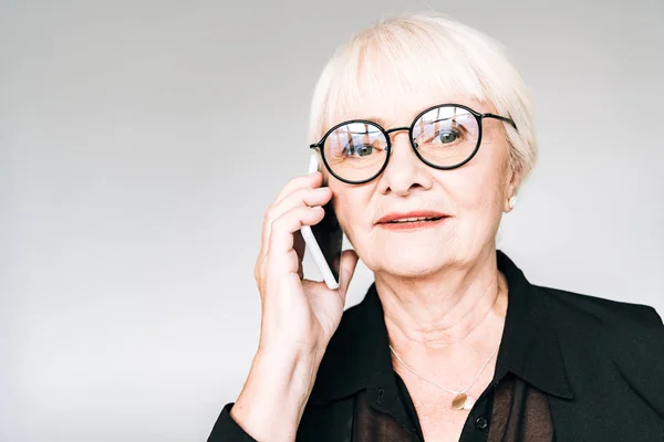 Senior businesswoman in black outfit and glasses talking on smartphone isolated on grey — Stock Photo