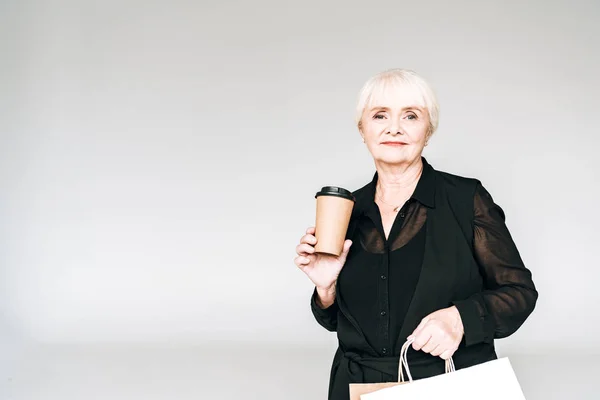 Mulher sênior elegante em roupa preta com sacos de compras e café para ir isolado em cinza — Fotografia de Stock