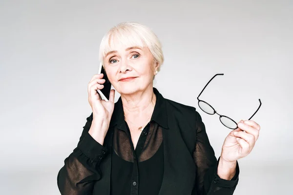 Senior businesswoman in black outfit with glasses talking on smartphone isolated on grey — Stock Photo