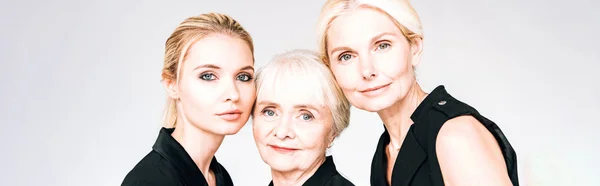 Plano panorámico de mujeres rubias de tres generaciones en trajes negros totales aislados en gris - foto de stock