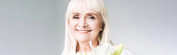 Plan panoramique de heureuse femme âgée blonde en vêtements blancs tenant des fleurs isolées sur gris — Photo de stock