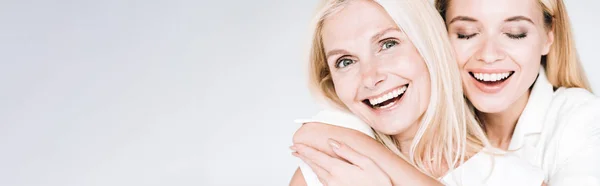 Panoramic shot of happy blonde mature mother and young daughter in total white clothes embracing isolated on grey — Stock Photo