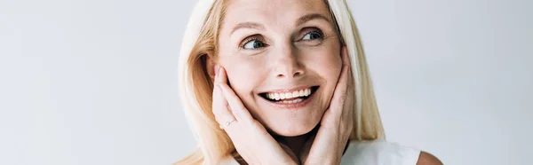 Panoramaaufnahme einer glücklichen blonden reifen Frau mit Händen in der Nähe des Gesichts, die isoliert von grau wegschaut — Stockfoto