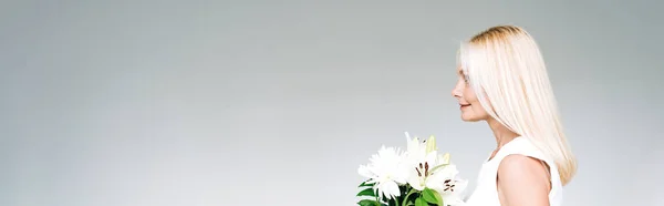 Вид сбоку счастливой блондинки зрелого возраста с белыми цветами, изолированными на сером, панорамном снимке — стоковое фото