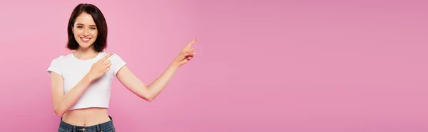 Panoramic shot of beautiful smiling girl pointing with fingers aside isolated on pink — Stock Photo
