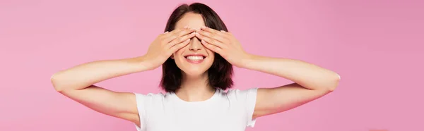 Tiro panorâmico de bela menina sorridente com as mãos em olhos isolados em rosa — Fotografia de Stock