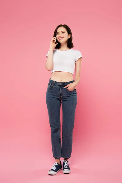 Vista de longitud completa de hermosa chica sonriente hablando en el teléfono inteligente aislado en rosa - foto de stock