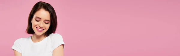Панорамний знімок красивої усміхненої сором'язливої дівчини, яка дивиться вниз ізольовано на рожевому — стокове фото
