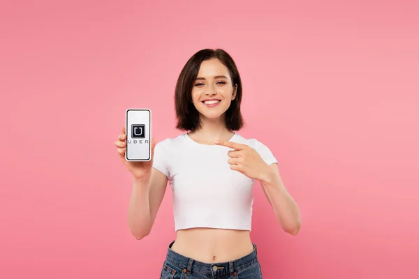 KYIV, UCRAINA - 3 LUGLIO 2019: ragazza sorridente che punta con il dito lo smartphone con logo uber isolato su rosa — Foto stock