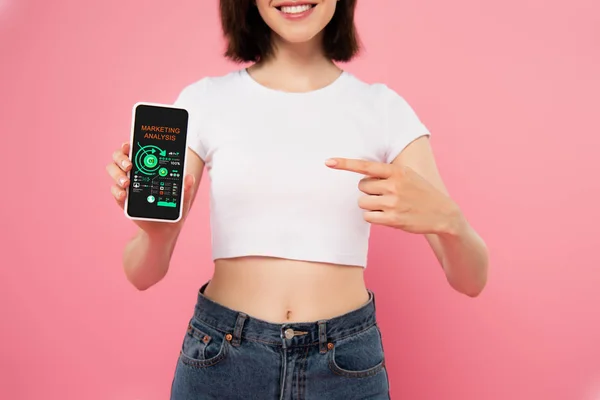 Recortado vista de chica apuntando con el dedo en el teléfono inteligente con aplicación de análisis de marketing aislado en rosa - foto de stock