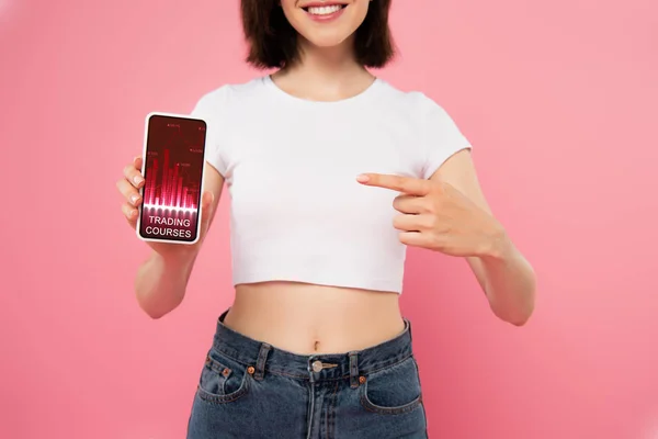 Recortado vista de chica apuntando con el dedo en el teléfono inteligente con cursos de comercio aplicación aislada en rosa - foto de stock