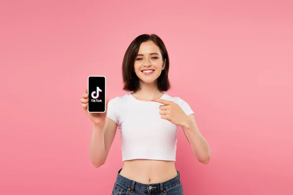 KYIV, UCRAINA - 3 LUGLIO 2019: ragazza sorridente che punta con il dito lo smartphone con logo tiktok isolato su rosa — Foto stock