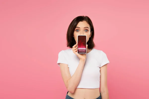 Surprised girl holding smartphone with trading courses app isolated on pink — Stock Photo