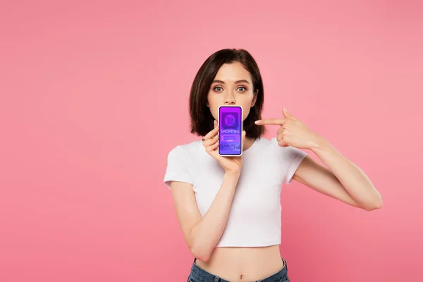 Chocado chica apuntando con el dedo en el teléfono inteligente con la aplicación de compras aislado en rosa - foto de stock