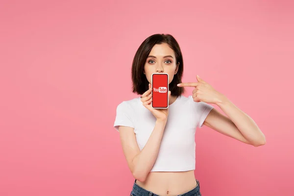 KYIV, UKRAINE - JULY 3, 2019: shocked girl pointing with finger at smartphone with youtube logo isolated on pink — Stock Photo