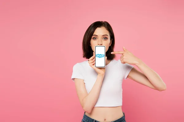 KYIV, UCRANIA - 3 de julio de 2019: chica impactada señalando con el dedo al teléfono inteligente con el logotipo de skype aislado en rosa - foto de stock