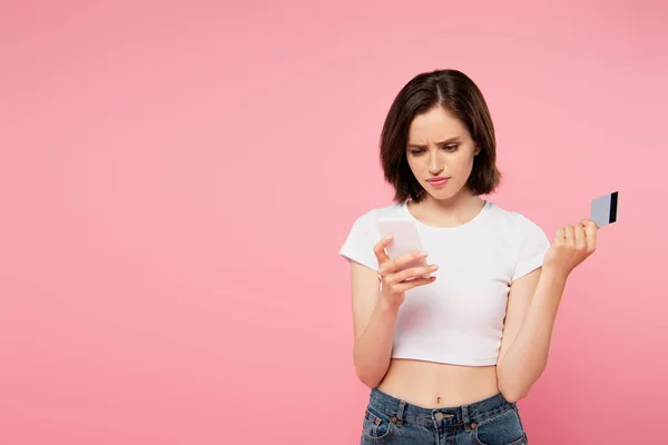 Chica confundida sosteniendo teléfono inteligente y tarjeta de crédito aislado en rosa - foto de stock