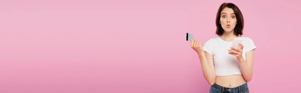 Plano panorámico de chica sorprendida sosteniendo teléfono inteligente y tarjeta de crédito aislado en rosa - foto de stock