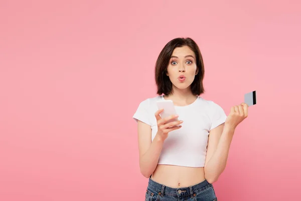 Chica sorprendida sosteniendo teléfono inteligente y tarjeta de crédito aislado en rosa - foto de stock
