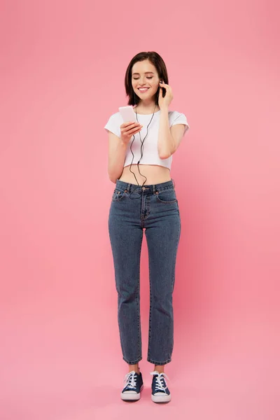 Vista completa de la chica sonriente escuchando música en auriculares con teléfono inteligente aislado en rosa - foto de stock