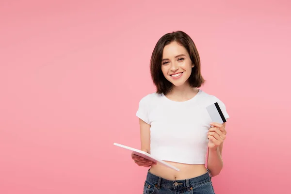 Улыбающаяся девушка держит цифровой планшет и кредитную карту изолированы на розовый — стоковое фото