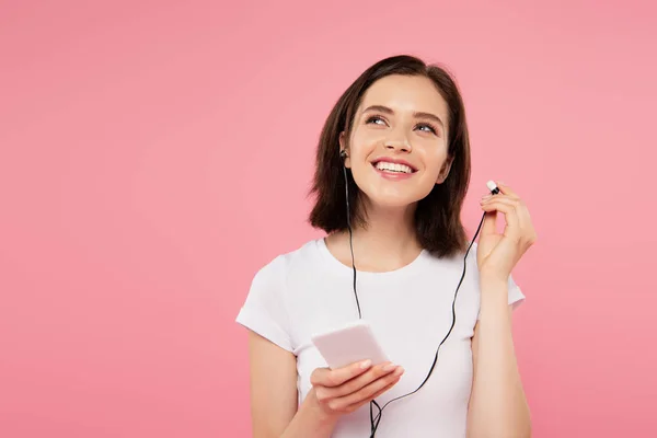 Chica sonriente soñadora escuchando música en auriculares con teléfono inteligente aislado en rosa - foto de stock