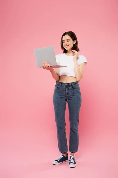 Vista de longitud completa de sonriente chica bonita sosteniendo portátil aislado en rosa - foto de stock