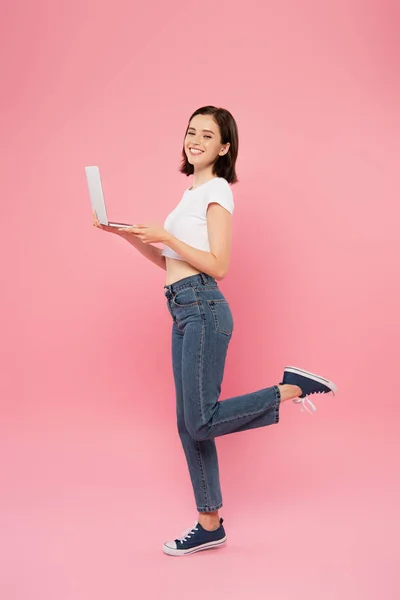 Vista completa de chica bonita sonriente posando con el ordenador portátil aislado en rosa - foto de stock