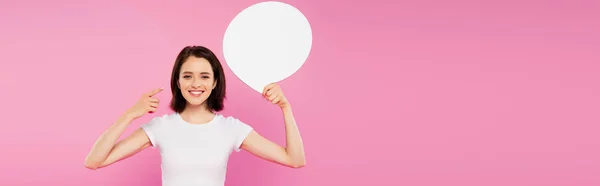 Panoramic shot of smiling pretty girl pointing with finger at blank white speech bubble isolated on pink — Stock Photo